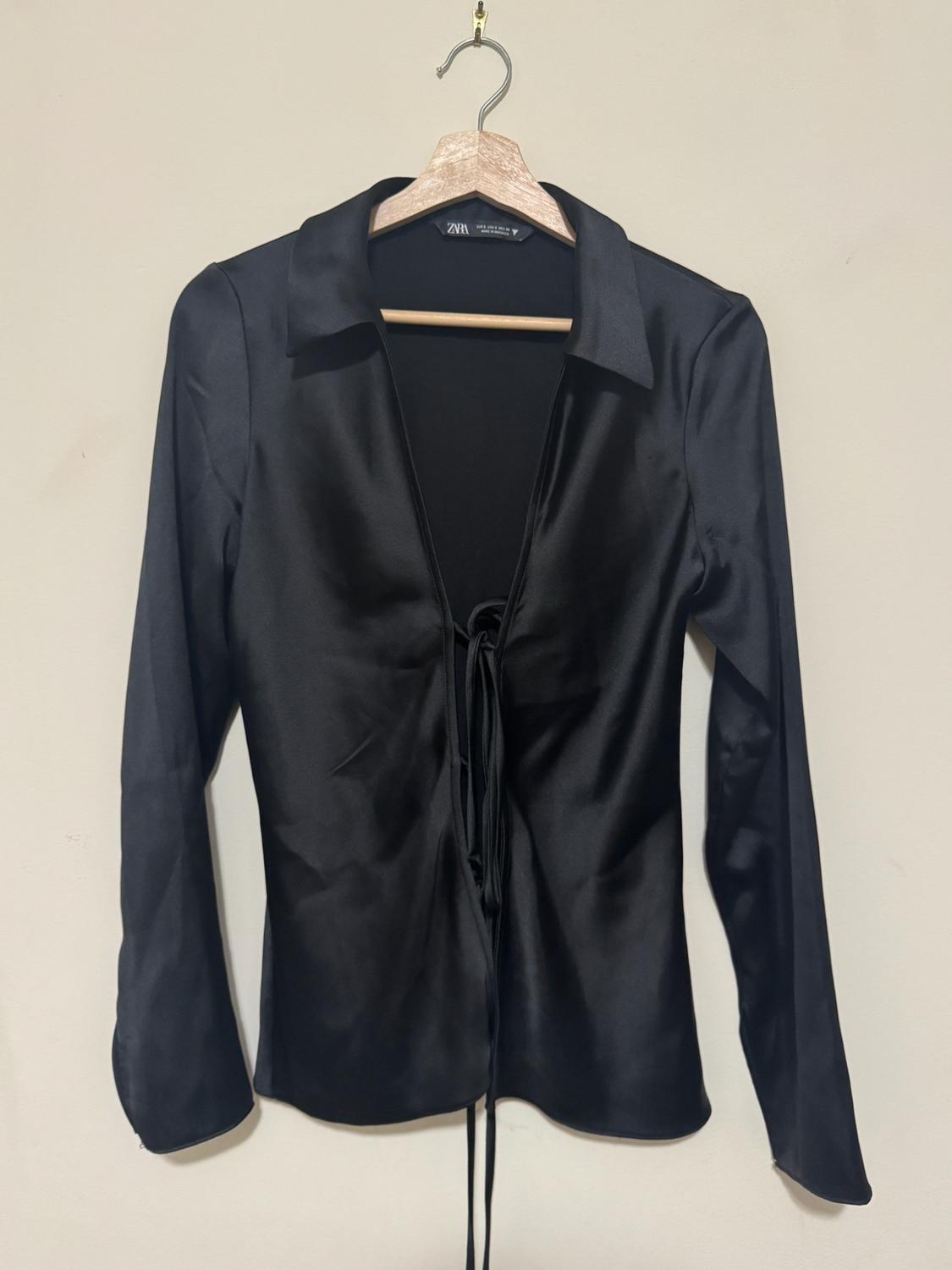Black long-sleeve blouse with a deep neckline and front lace-up detail, hanging on a wooden hanger against a light background. Text ZARA.