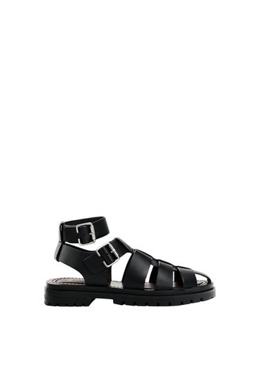 Black strappy sandals with metallic buckles and a thick sole, on a white background. Modern and minimalist design.