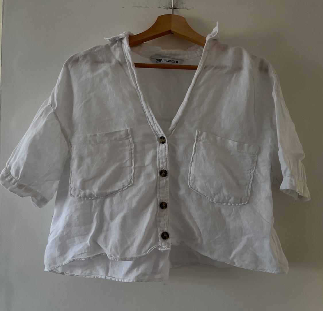 White short-sleeved shirt, V-neck, buttons and two front pockets, hanging on a wooden hanger against a white wall. ZARA label.