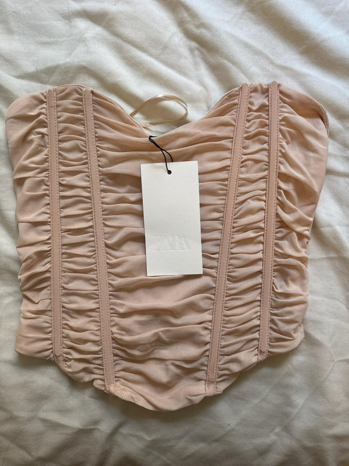 Nude corset-style top with ruched fabric and a label featuring the text ZARA, laid on a crumpled white fabric.