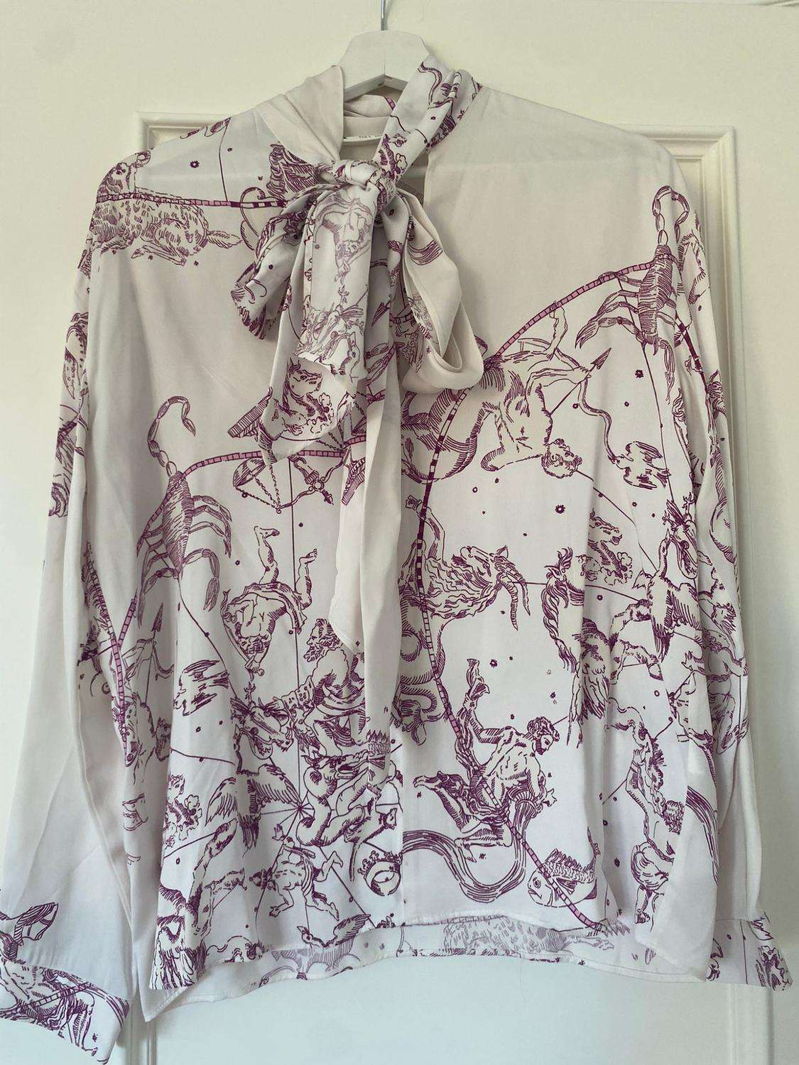 White blouse with an illustrated print in burgundy tones, a bow neck, hanging on a white hanger in front of a door.