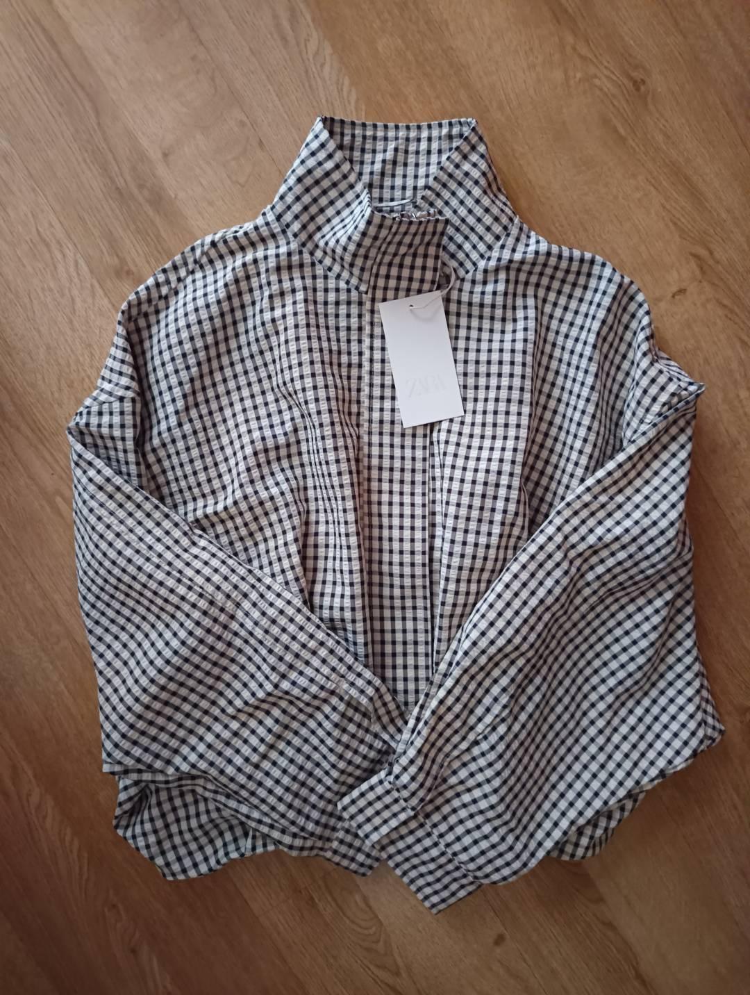 Black-and-white checked shirt with a folded high collar lying on a wooden floor. It has a label bearing the text ZARA.