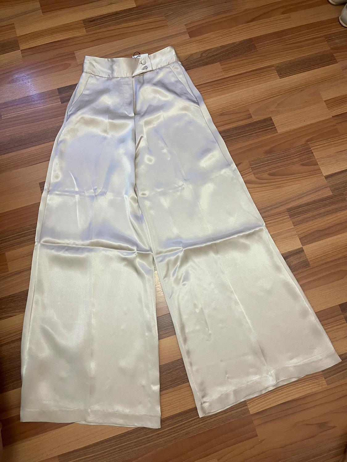 Cream wide-leg satin trousers with a high waist and buttons, on a light wooden floor.