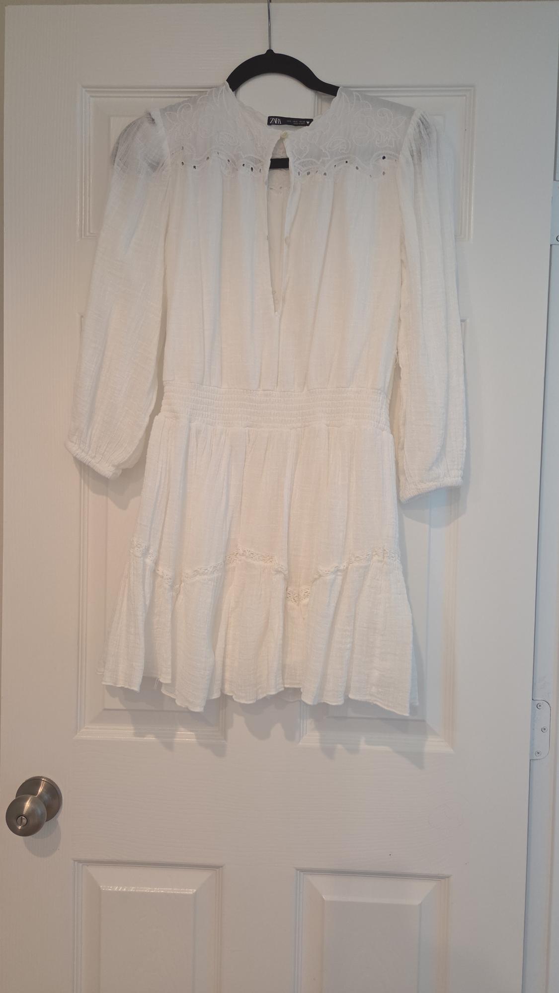 Long-sleeved white blouse with embroidered details on the collar, hanging on a black hanger over a white door.