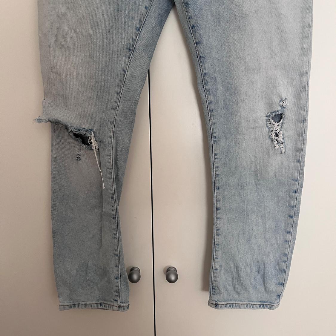Light blue distressed denim jeans with rips on both legs, hanging in front of a white door.