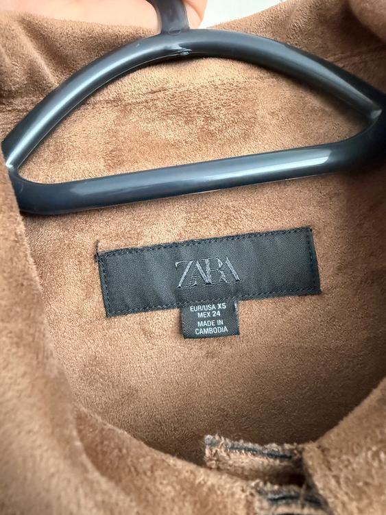 Brown jacket label on a black hanger. Visible text: "ZARA, EUR/USA XS, MEX 24, MADE IN CAMBODIA". Soft-looking fabric.