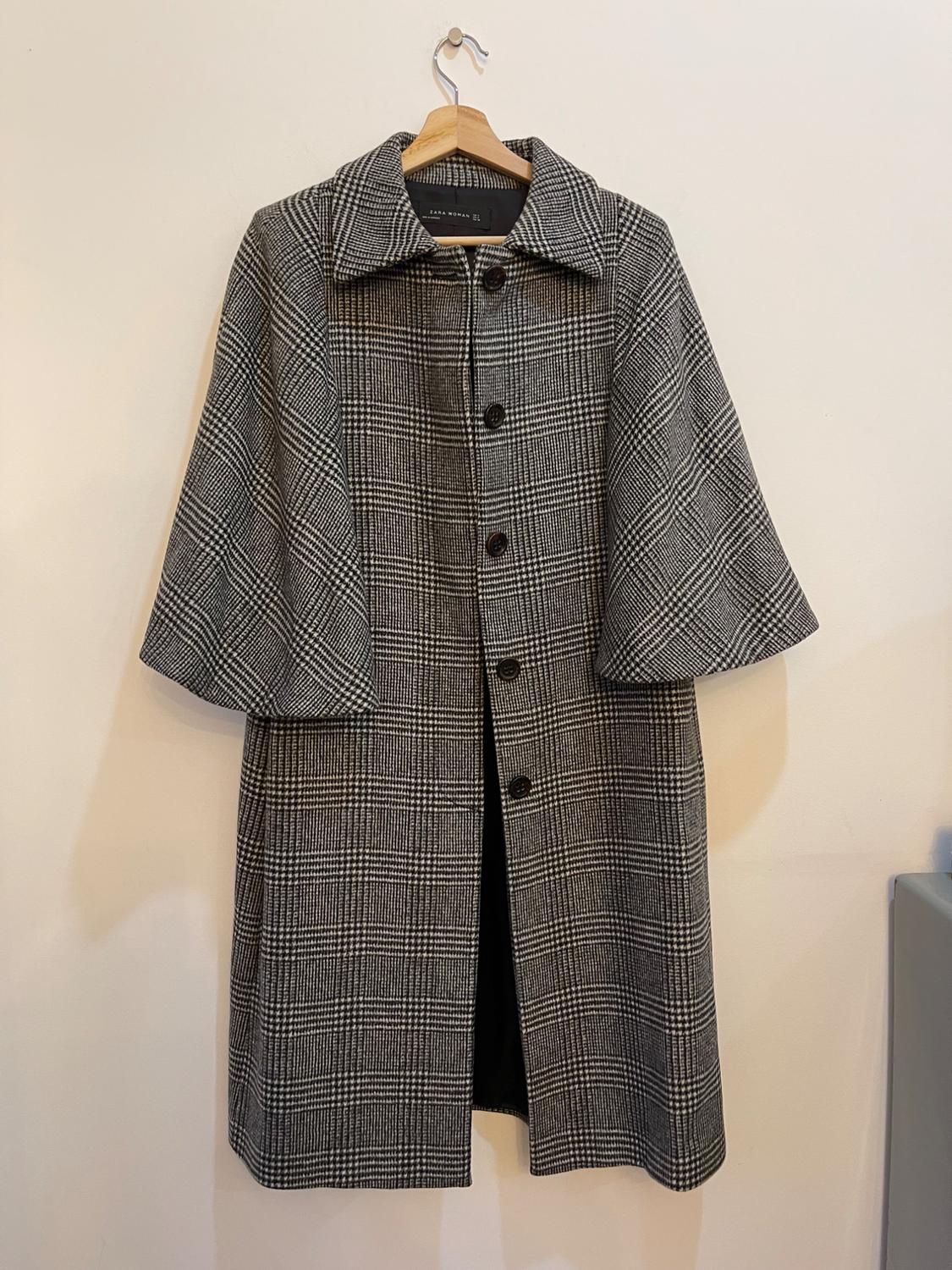 Grey checked coat with black buttons hanging on a wooden hanger against a white wall.