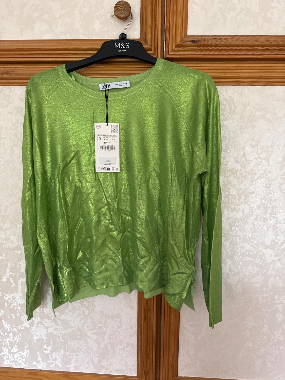 Bright green jumper hanging on a black M&S hanger, with visible price tag. Light textured wall background and wooden frame.