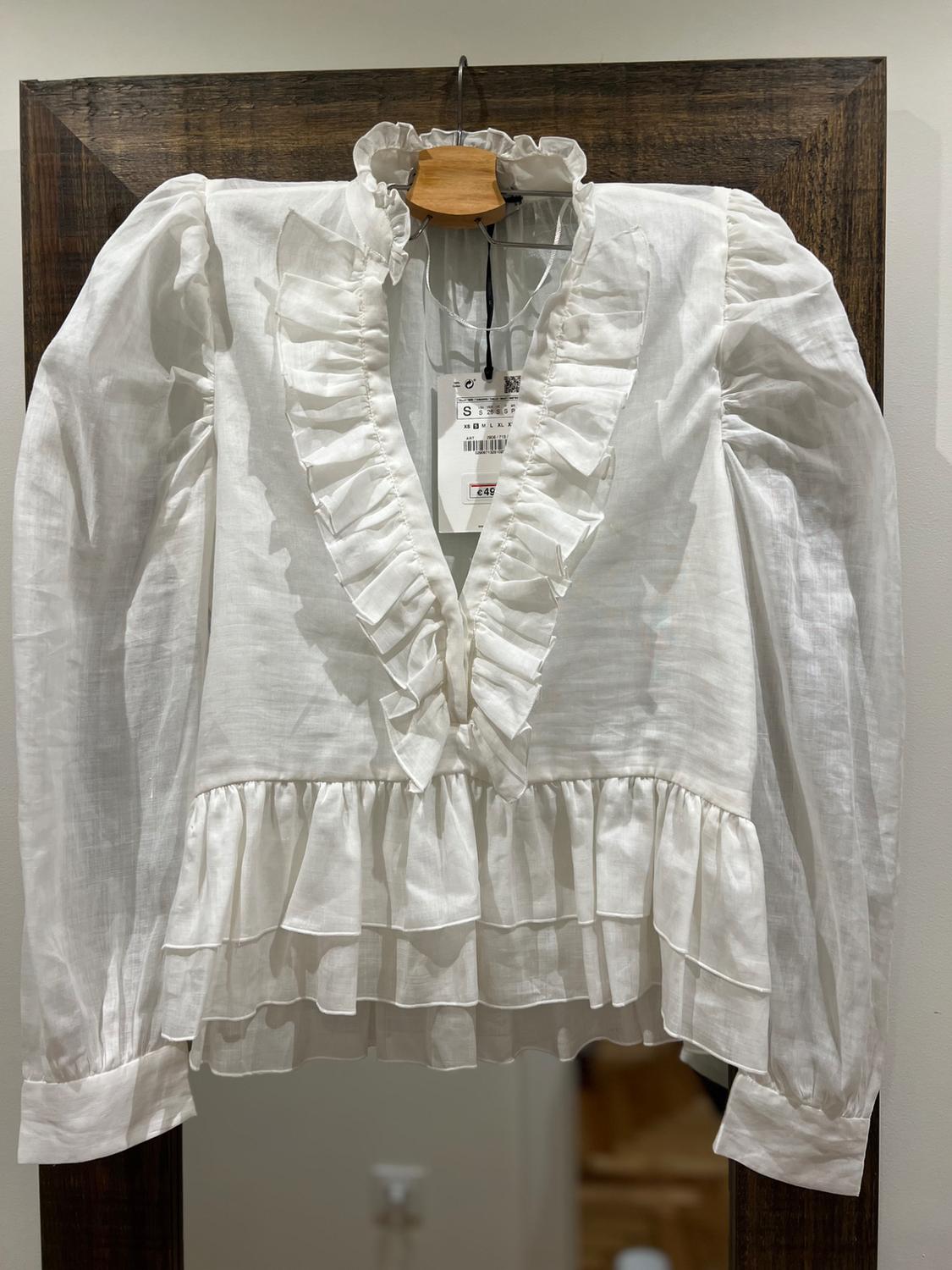 White blouse with ruffles on the collar and front, hanging on a wooden hanger. Visible tag with price of €49.