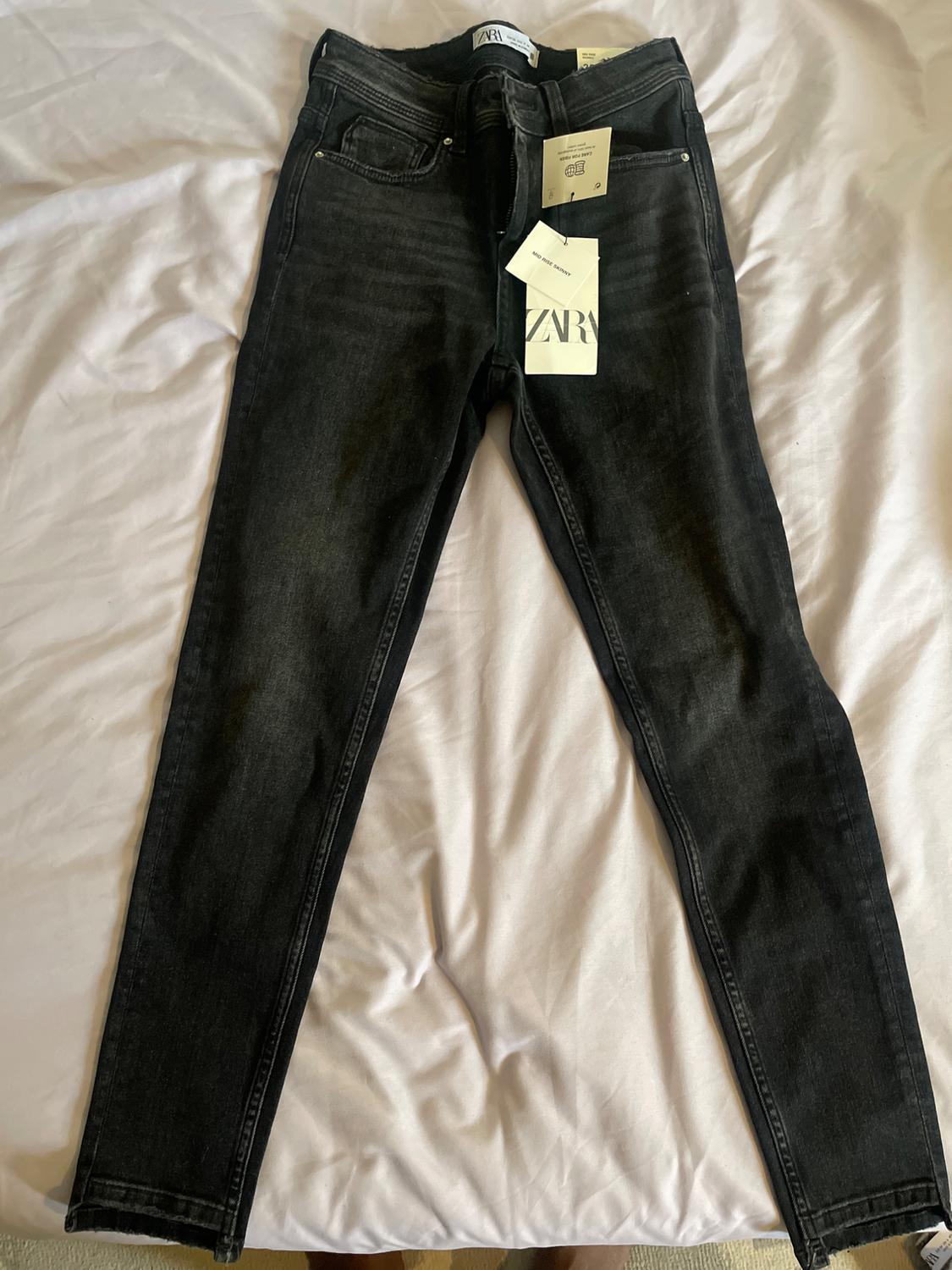 Black jeans on a white bed, with tags hanging that include the text "ZARA".