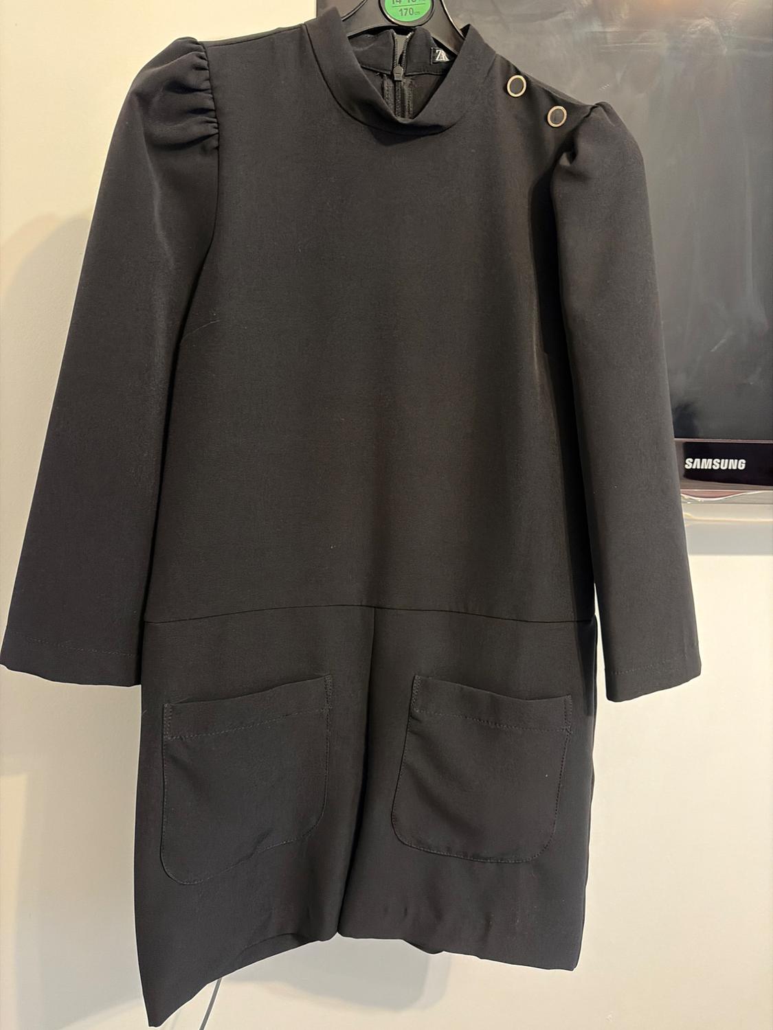 Black dress with long sleeves and shoulder pads, two front pockets and eyelet details on the shoulder. Light background and Samsung screen.