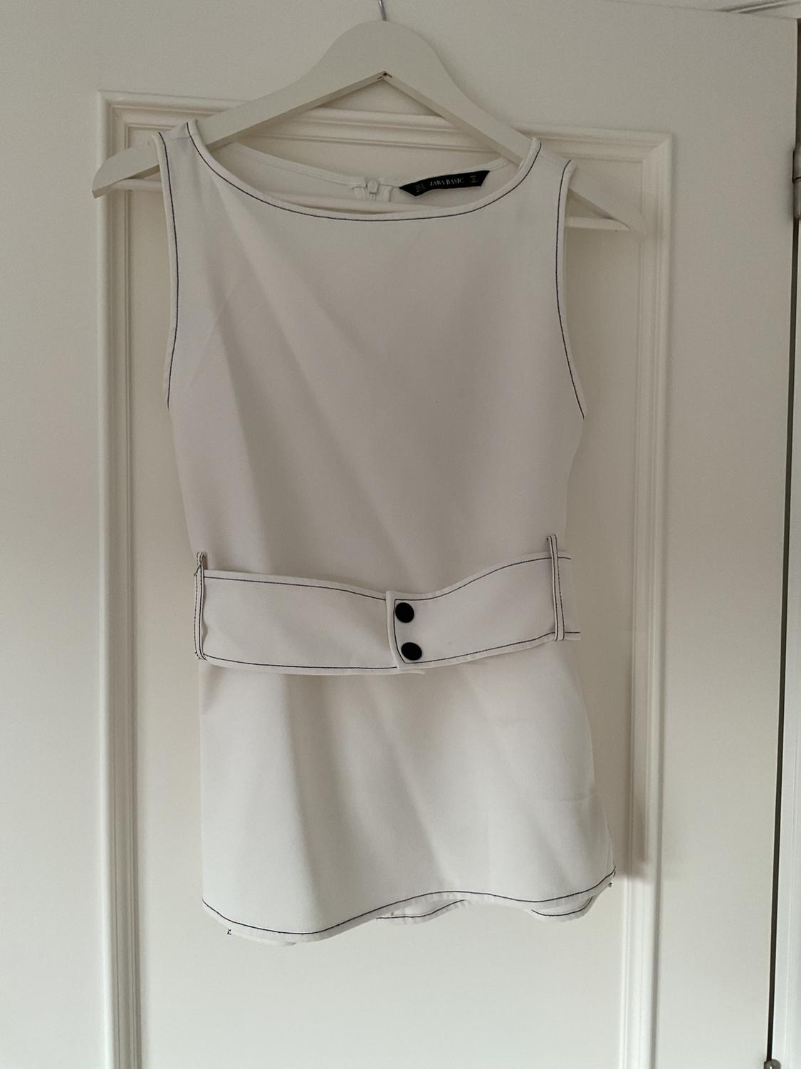 White sleeveless blouse with a wide belt and black buttons, hanging on a white hanger over a white door.