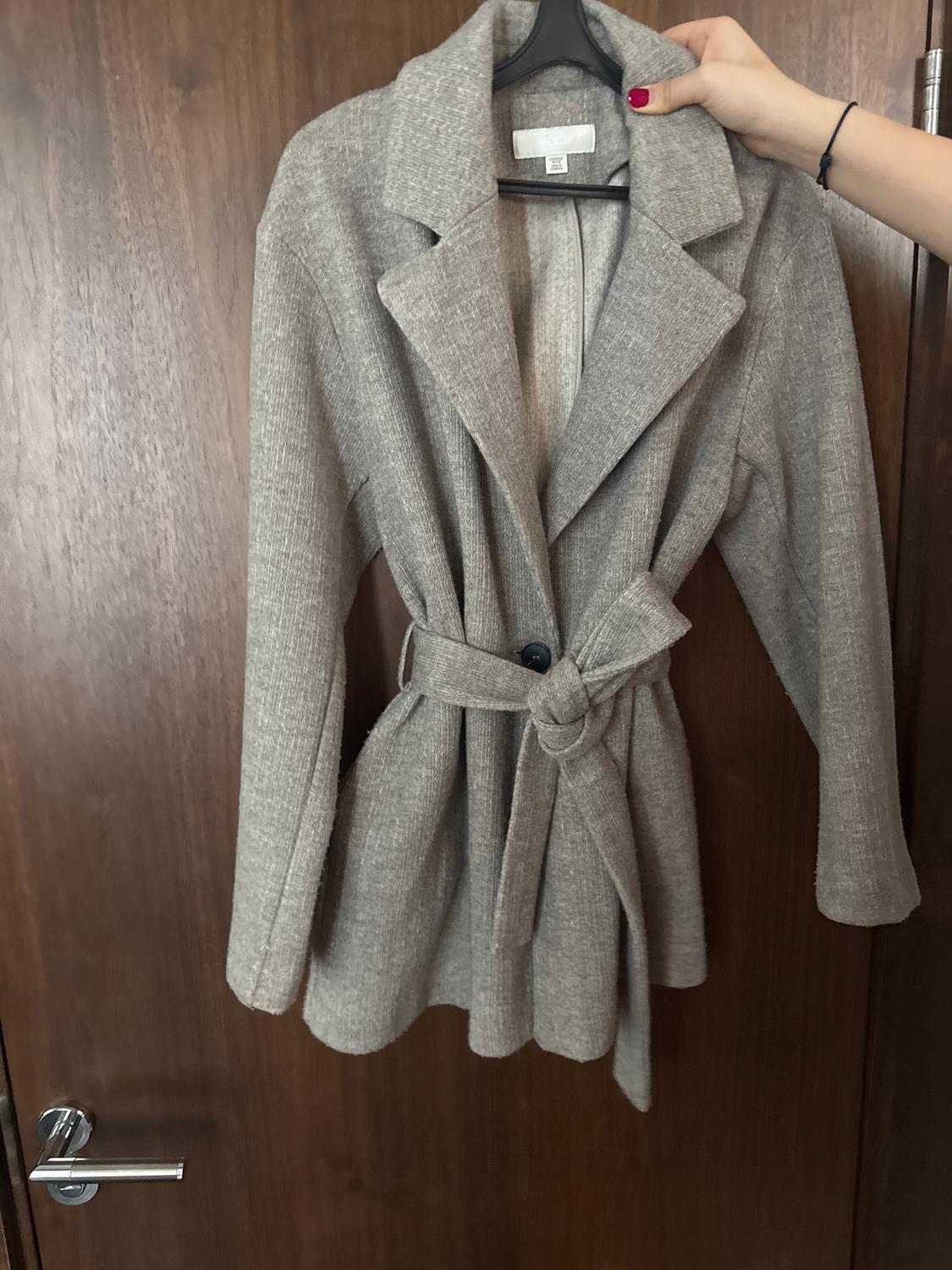 A grey, textured fabric coat is hanging on a black hanger, held by a hand with red painted nails, over a wooden door.