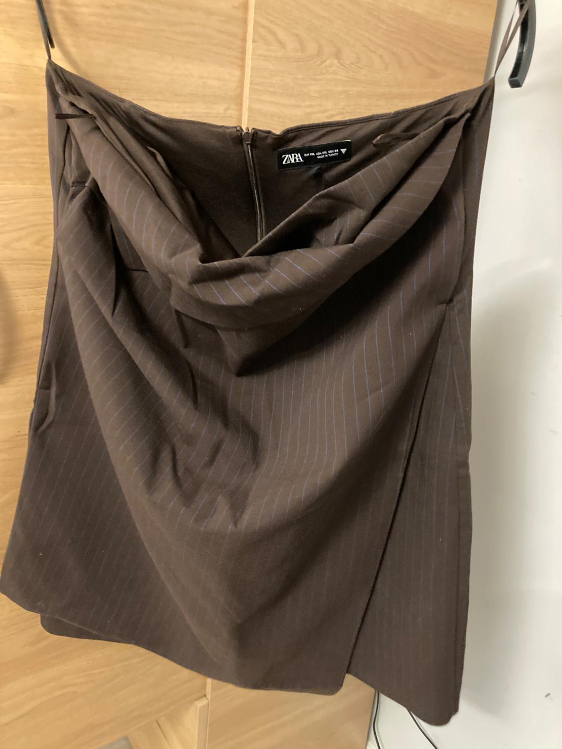 Brown pleated skirt with zip fastening, hanging on a hanger. Label visible with the text "ZARA". Light wood background.