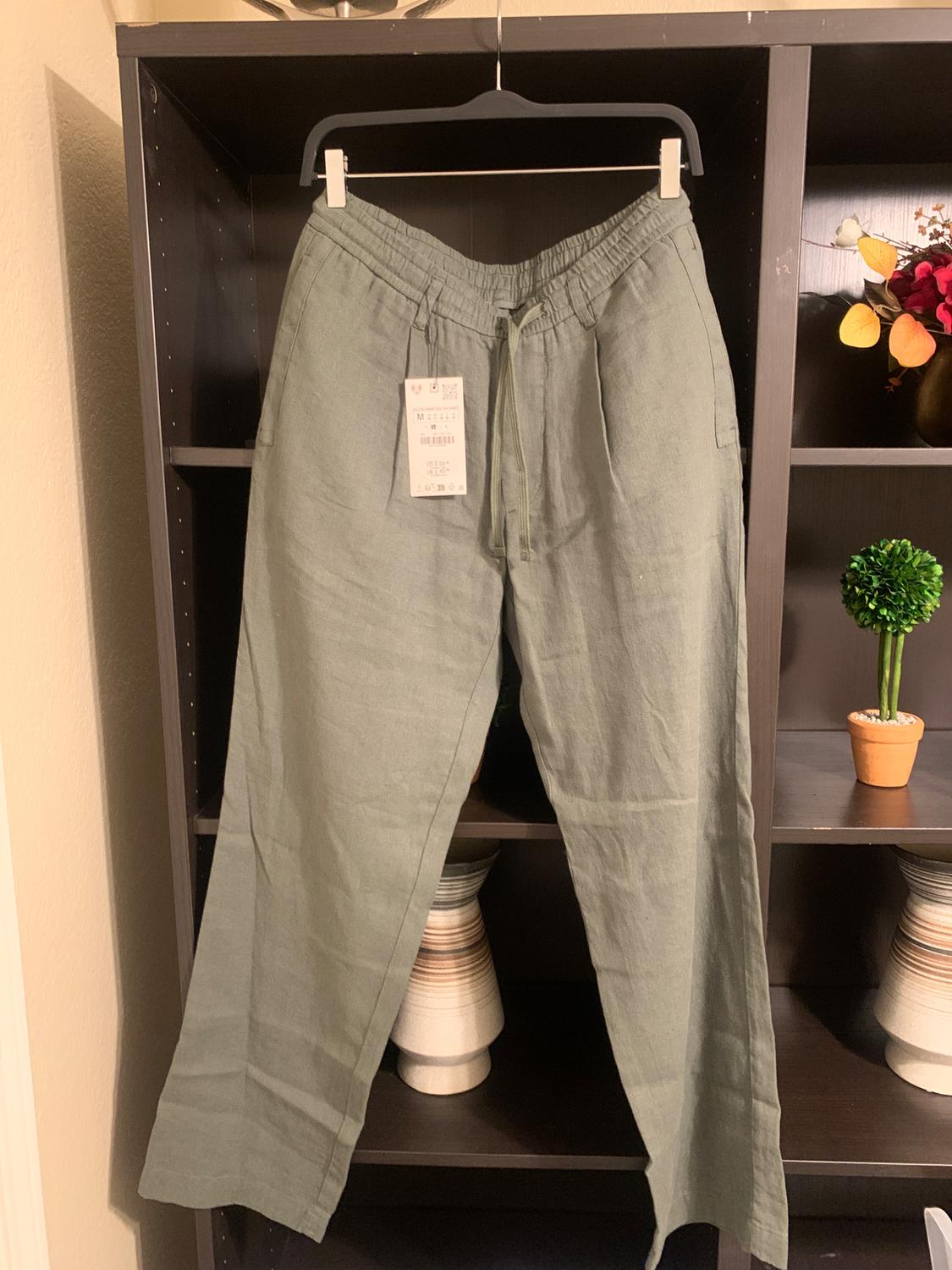Green linen trousers hanging on a hanger in front of a dark shelf, with a visible label. Plant and vase decorations around.