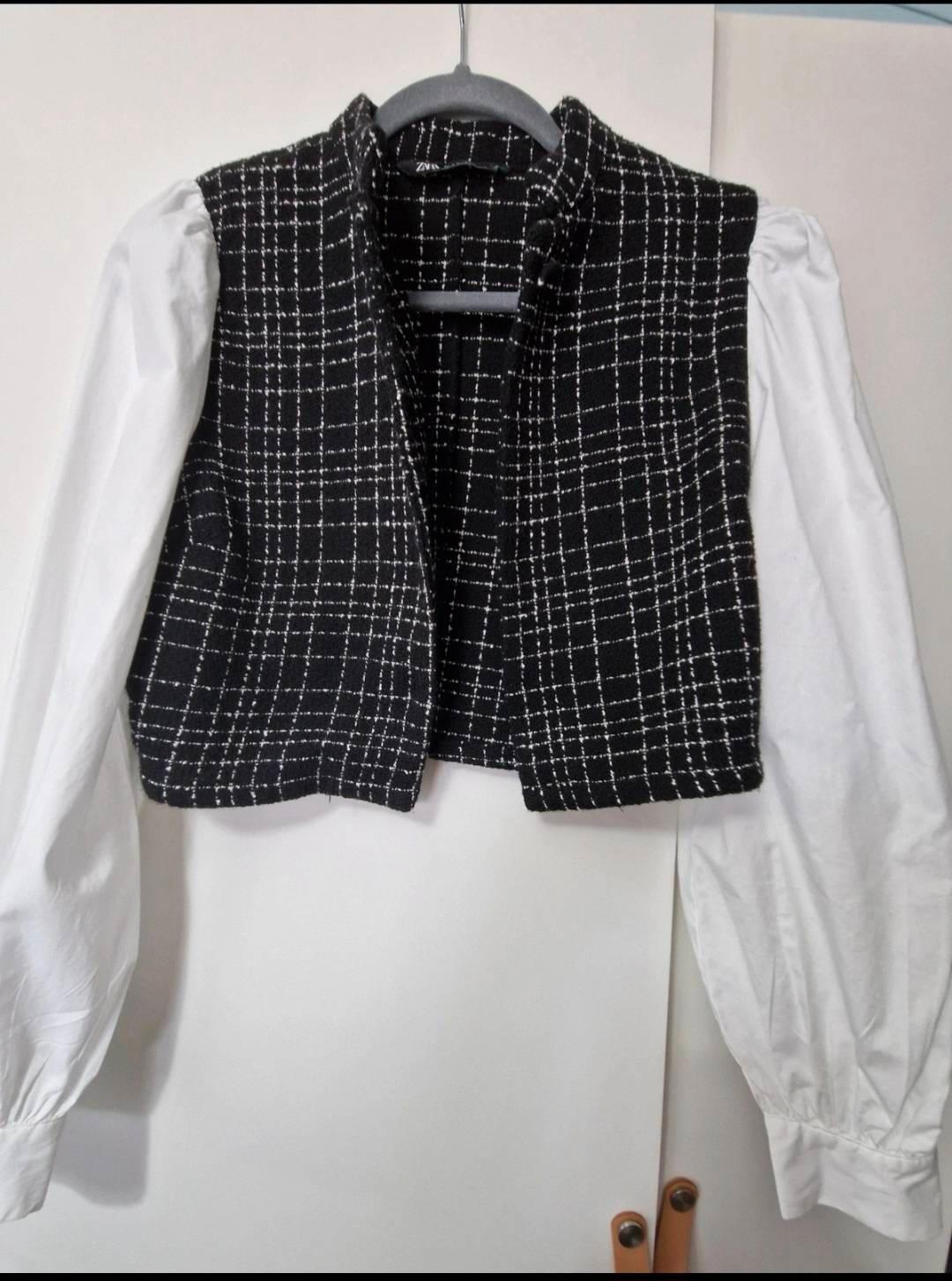 Black waistcoat with a white check pattern over a long-sleeved white shirt, hanging on a hanger in front of a white door.