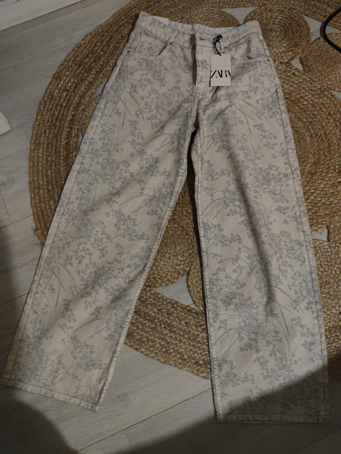Light grey floral print denim trousers on a jute rug. Visible label with text "ZARA".