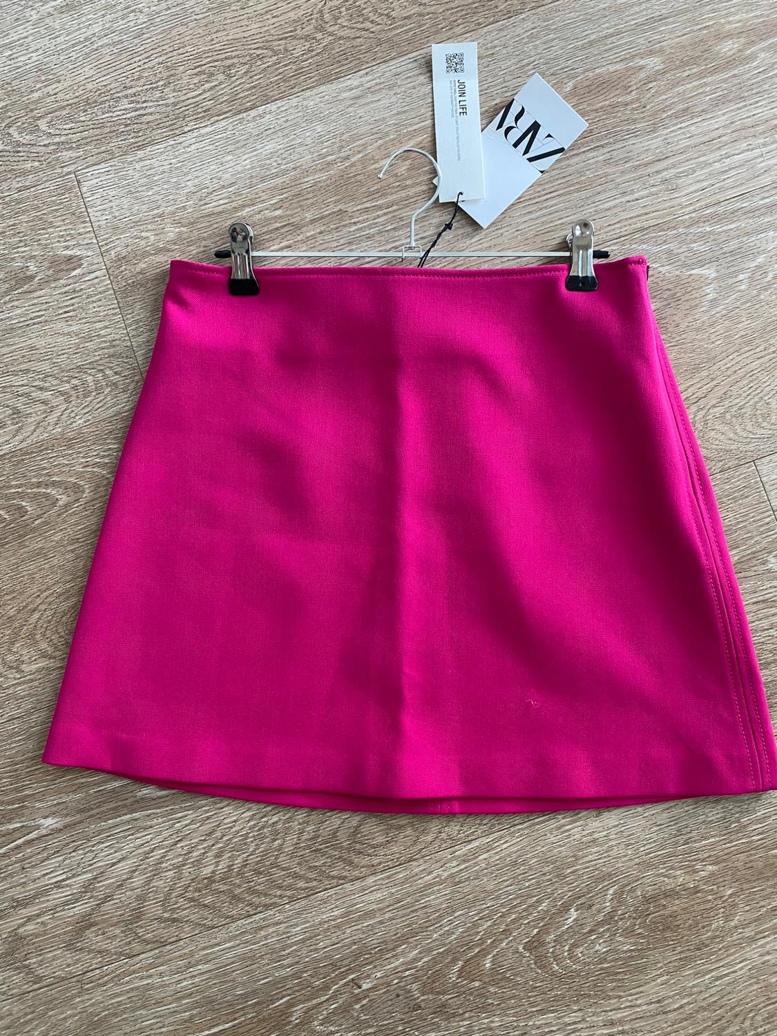 Fuchsia miniskirt hanging on a hanger over a light wood floor. Visible tags with text "JOIN LIFE" and "ZARA".