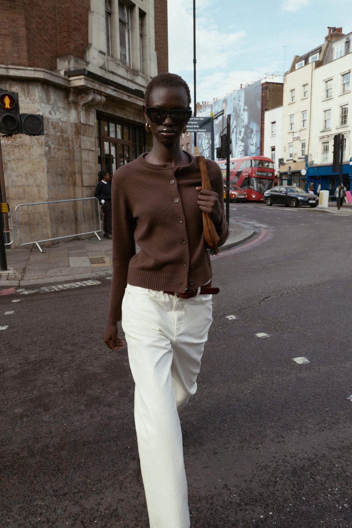 Komplet rjave pletene jakne z okroglim ovratnikom in gumbi ter belih ravnih hlač.