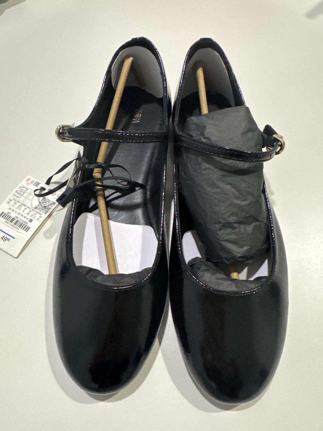 Black patent leather shoes with strap and buckle, stuffed with paper and wooden support. Barcode label visible on the left.