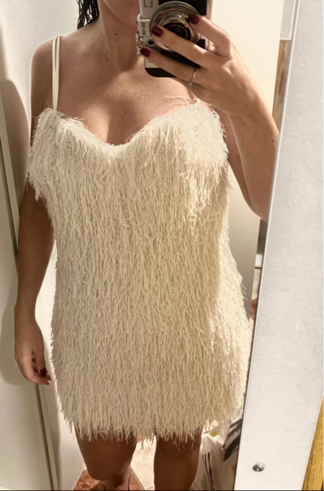 Woman in a changing room wearing a short, cream, fluffy-textured dress, holding a camera. Dark red painted nails.