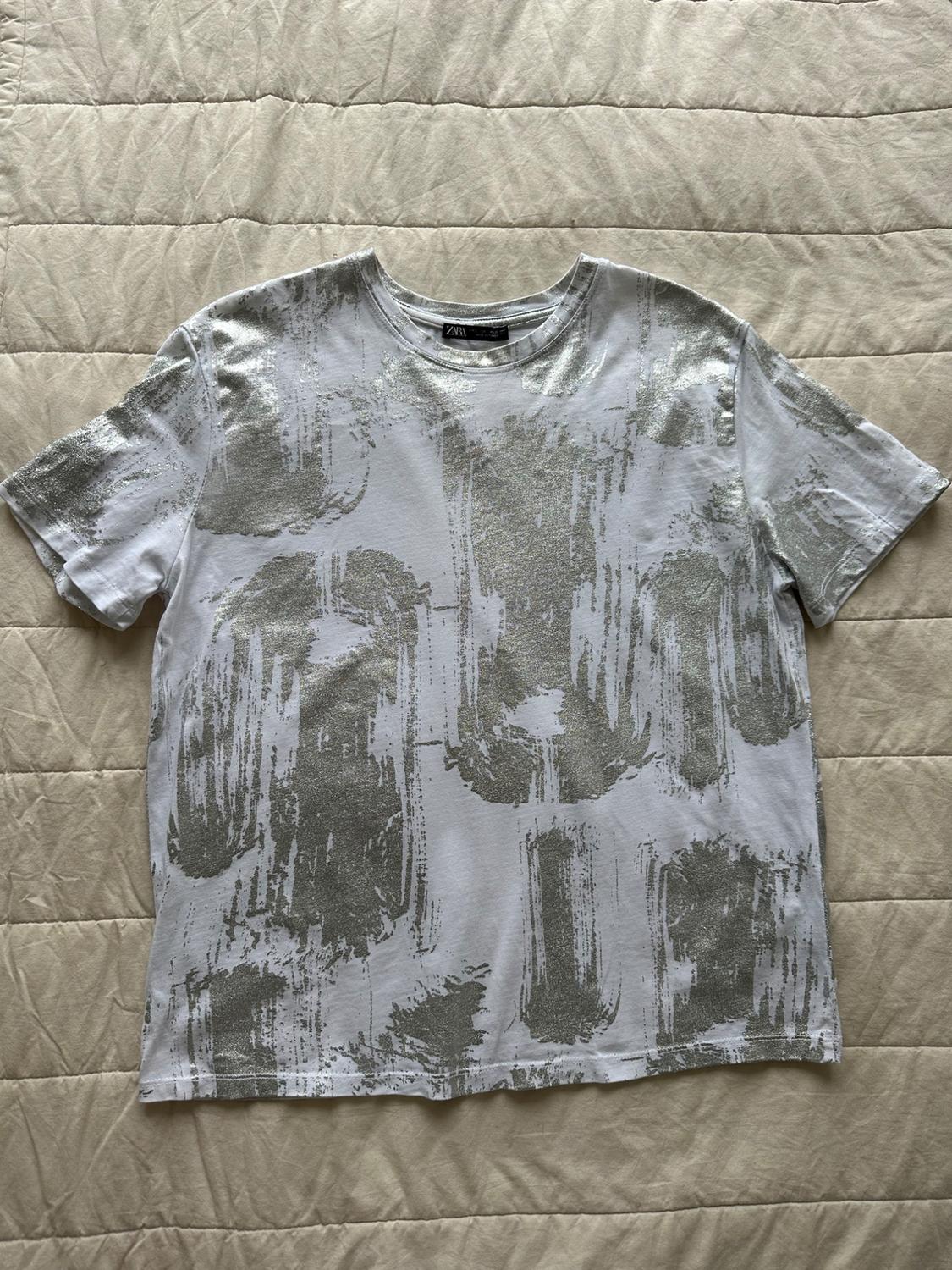 White short-sleeved T-shirt with a silver brushstroke print on a beige quilt. ZARA is visible on the neck label.