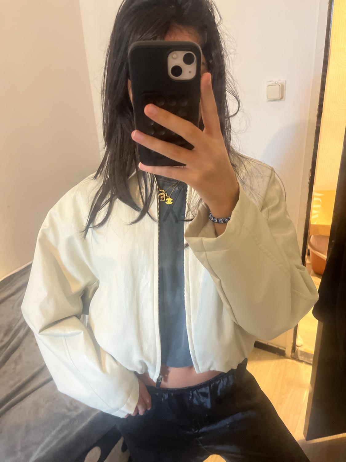 Mirror selfie of a person with their mobile phone obscuring their face. They are wearing a white jacket, dark T-shirt, black trousers and a gold necklace.