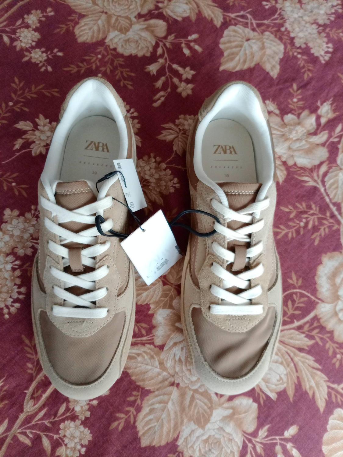 Pair of beige trainers with white laces against a maroon floral background. The insoles read ZARA SNEAKERS 39.