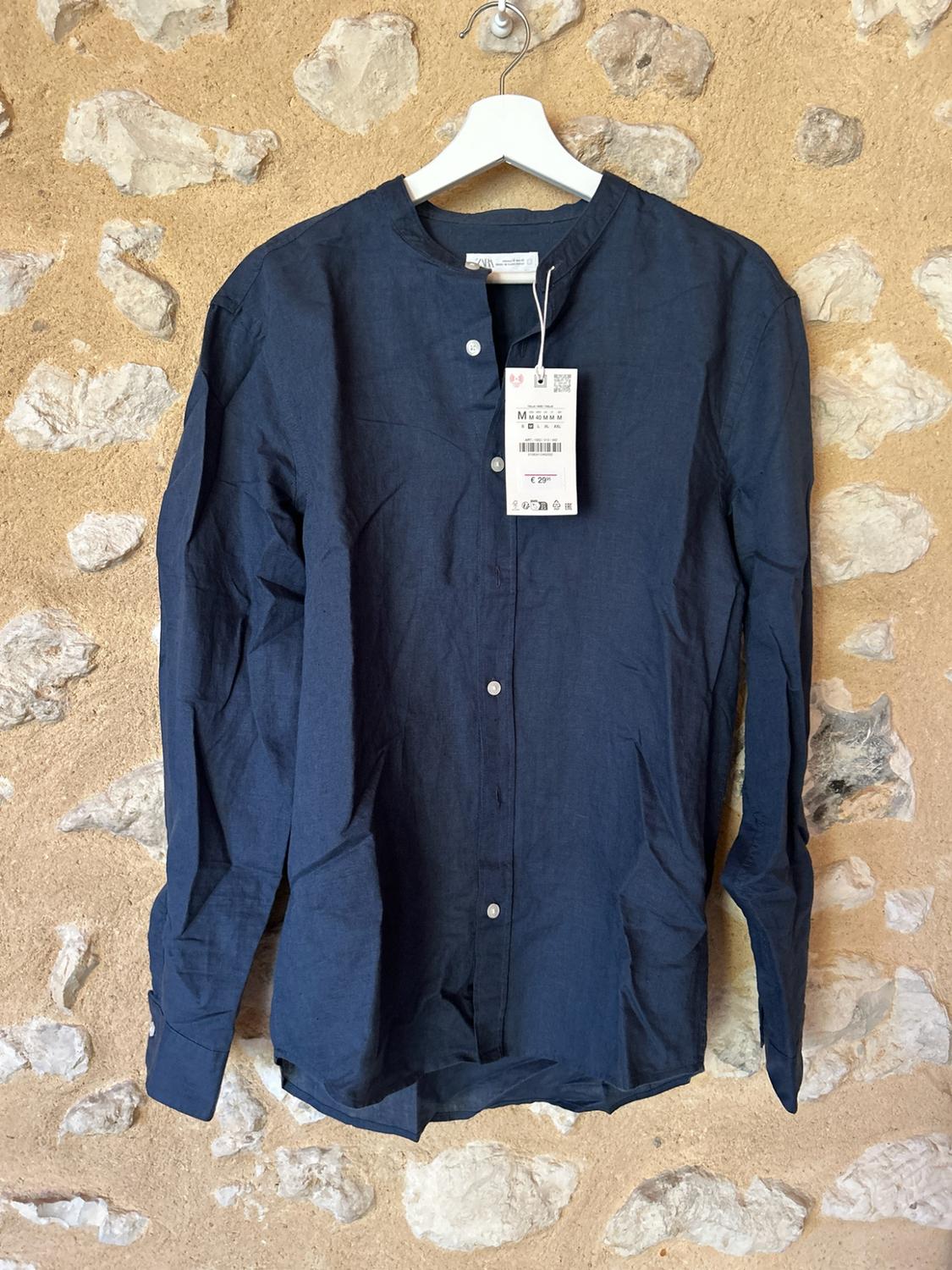 Navy blue long-sleeved shirt with a mandarin collar and label, hanging on a white coat hanger in front of a stone wall.