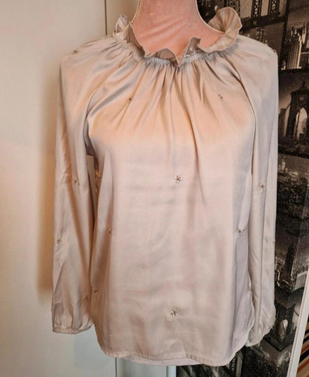 Beige blouse with a ruffled collar and small embroidered floral details, displayed on a mannequin in front of a decorative background.