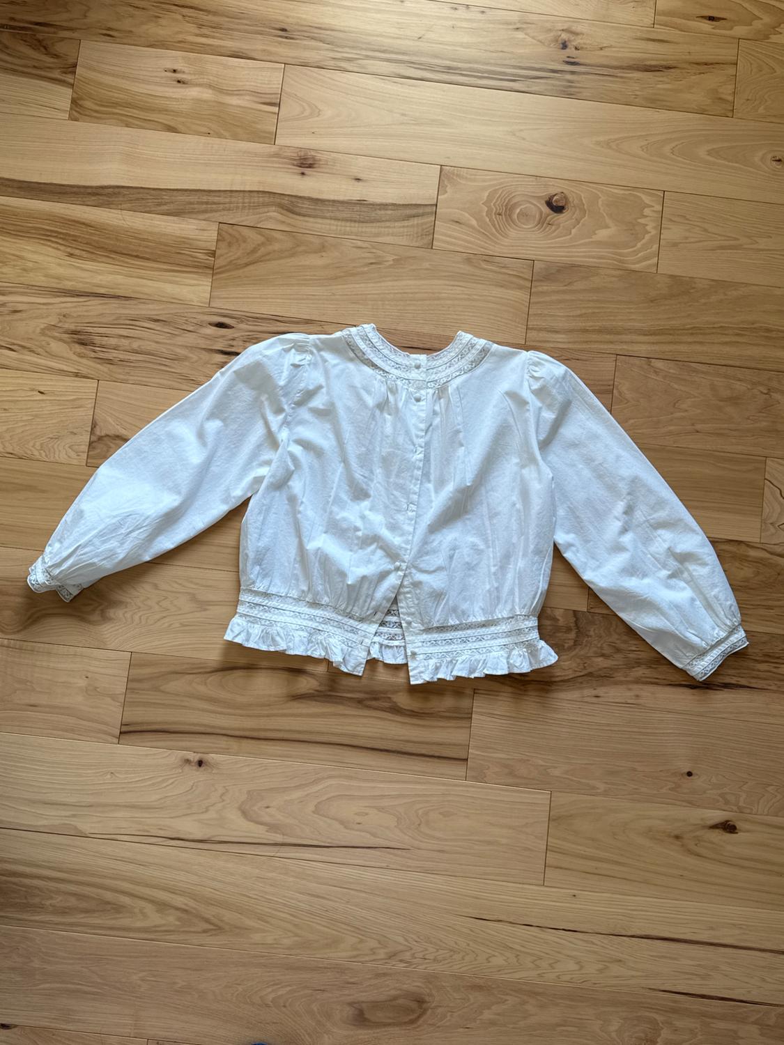 White long-sleeved blouse with lace detailing on the collar, cuffs, and waist, on a light wood floor.