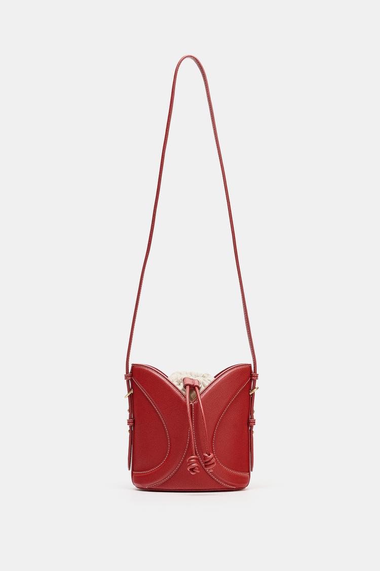 Přední pohled na červenou kabelku se stahovací šňůrkou a zakřivenými detaily. Dlouhý nastavitelný popruh a viditelné prošívání.