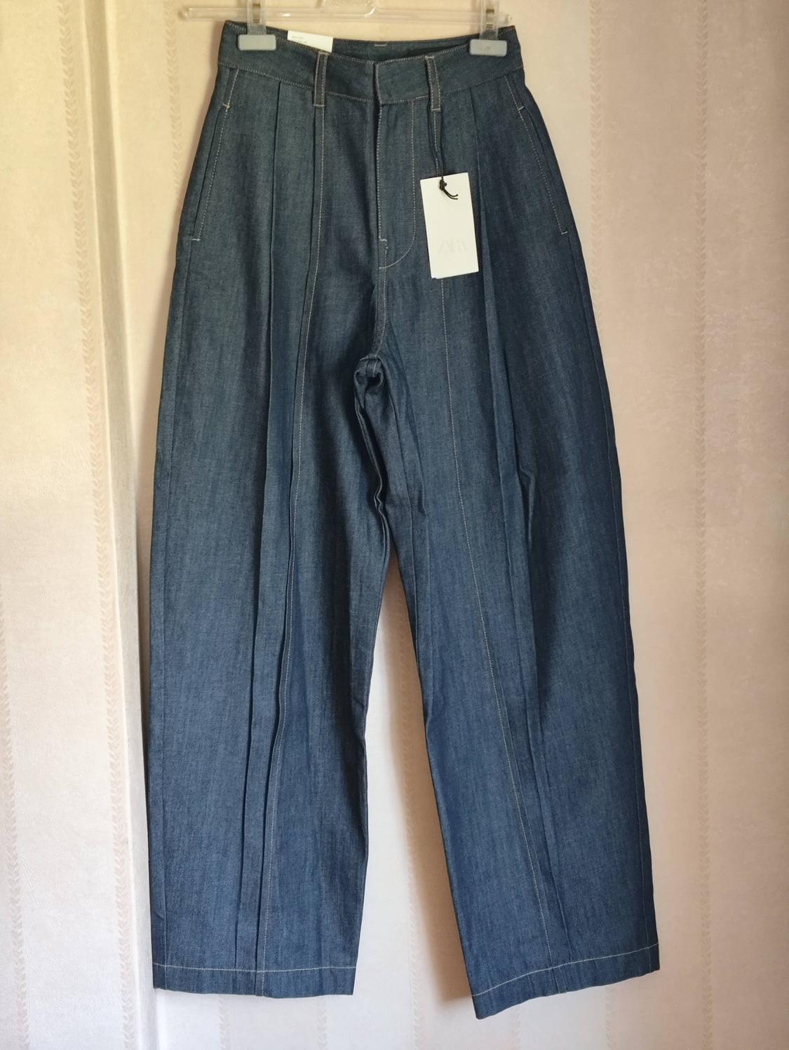 High-waisted blue jeans with front seams, hanging on a hanger against a light-coloured wall. Label with the text ZARA.