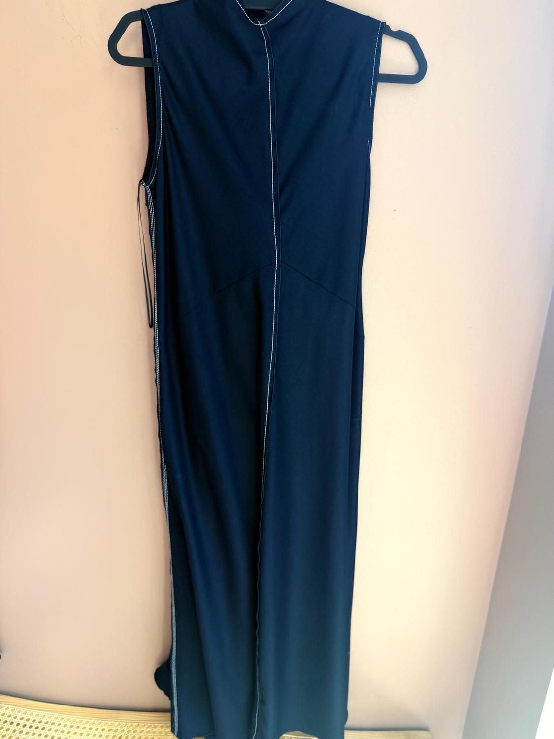 Long navy blue sleeveless dress with contrasting white stitching, hanging on a black hanger in front of a light-coloured wall.