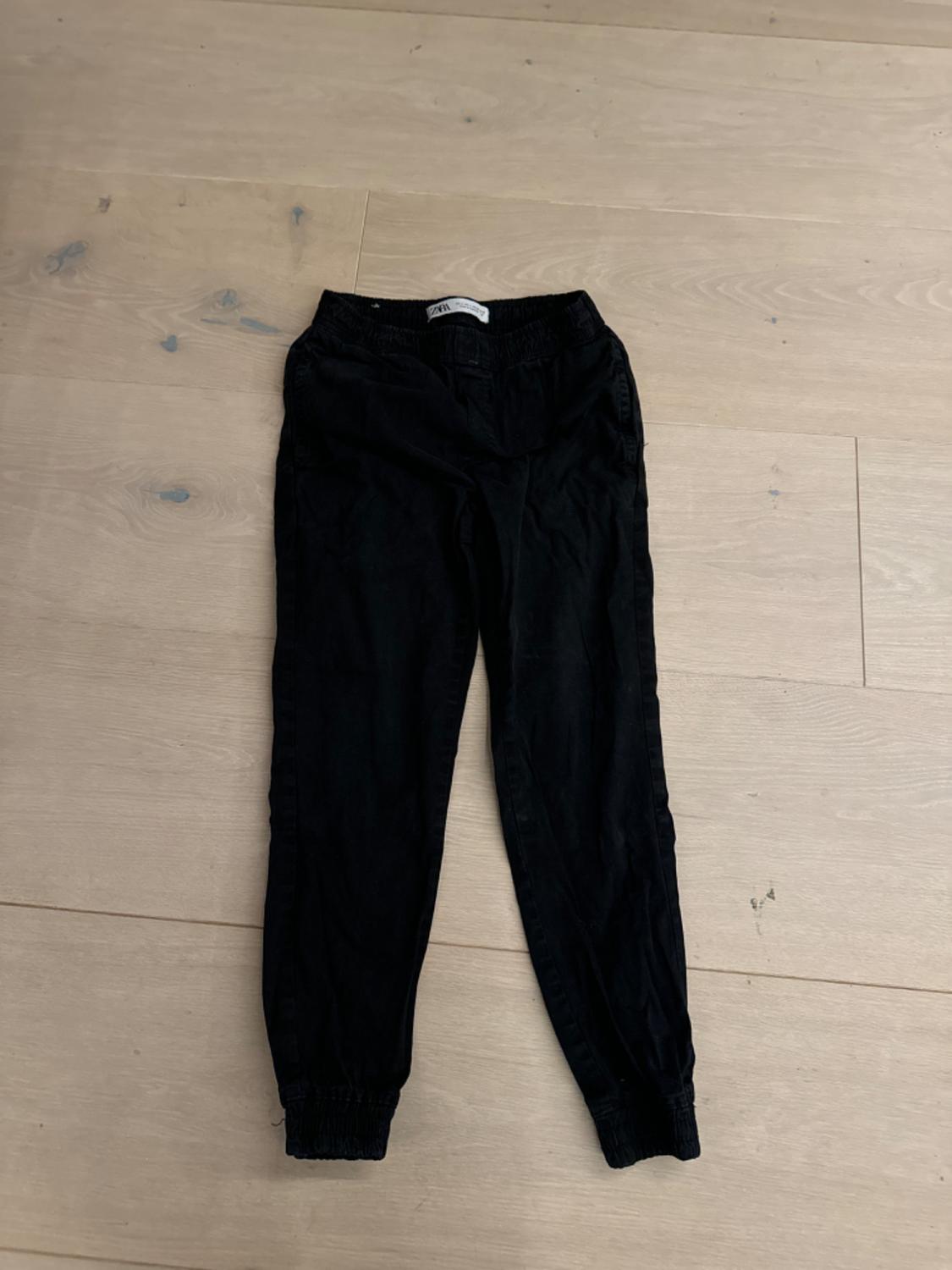 Black trousers made of soft fabric with an elasticated waist and fitted ankles, on a light wooden floor.