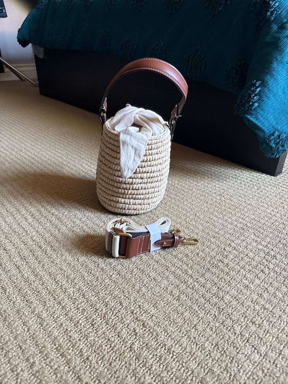 Woven basket bag with a brown leather handle and fabric lining, alongside a detachable strap, on a beige rug in front of a bed.
