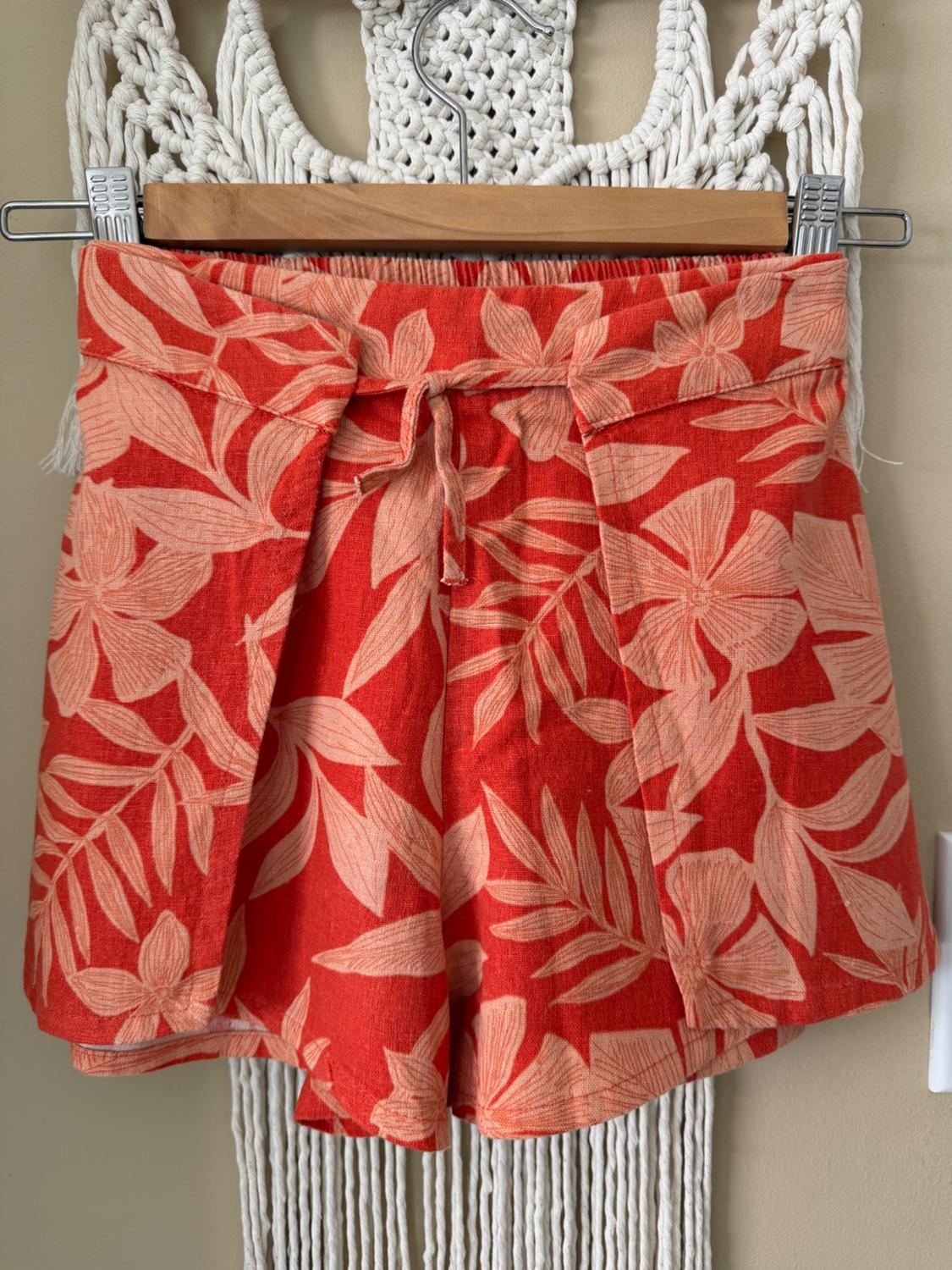 Red shorts with a beige leaf pattern hanging on a wooden hanger against a white macrame background.