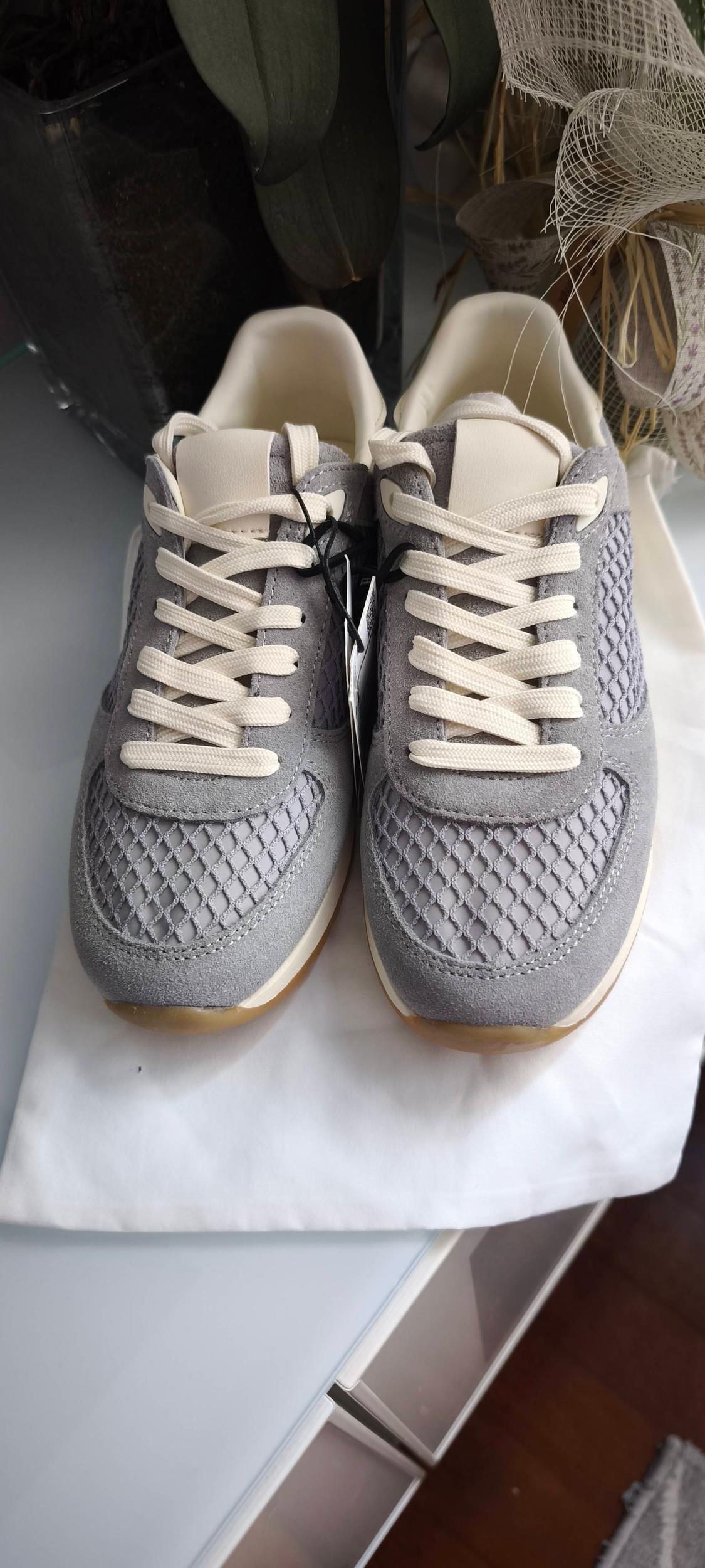 Grey trainers with white laces on a white surface, mesh and suede details, background with decorative plants.