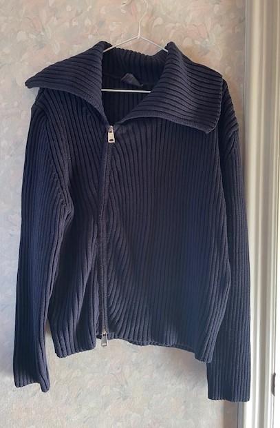 Dark blue knitted jacket with a zip and a wide collar, hanging on a hanger against a light textured background.
