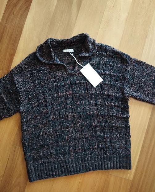 Chunky knit jumper in dark tones with a high neck, white hanging tag, on a light wooden floor.