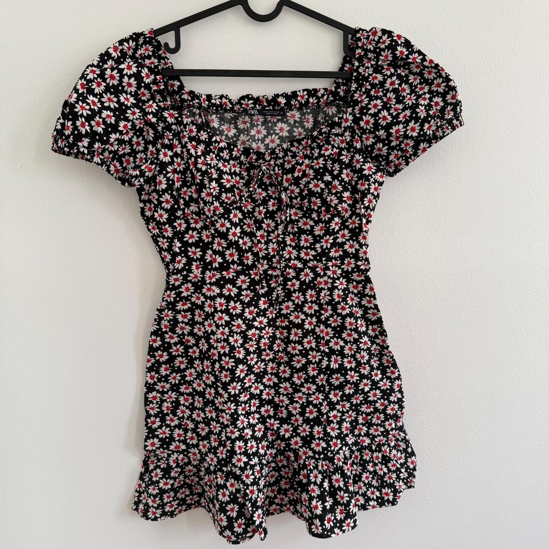 Short black dress with a white daisy print and red details, puffy sleeves, and a gathered neckline, hanging on a black hanger.