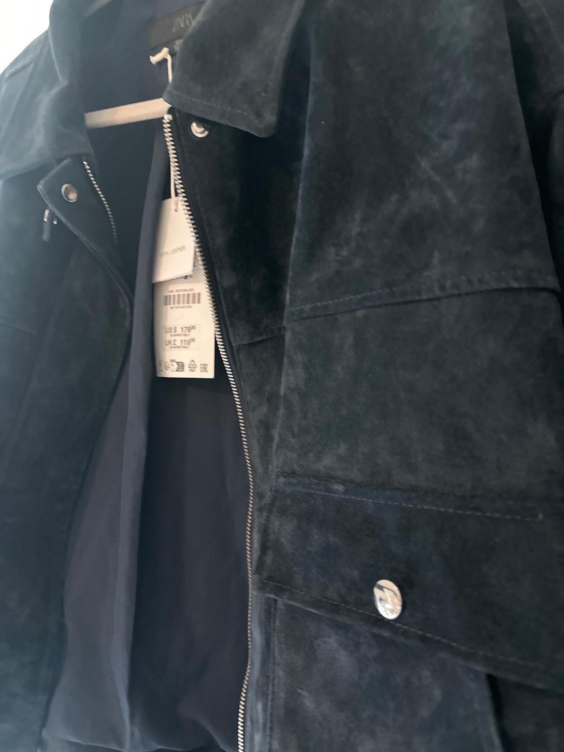 Black suede jacket hanging on a hanger, with an open zipper and visible tags inside.