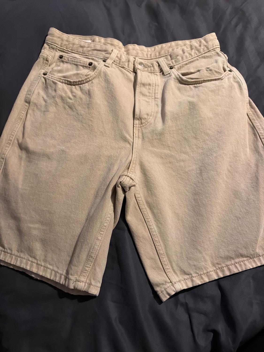 Beige five-pocket denim shorts, on a background of dark crinkled fabric.