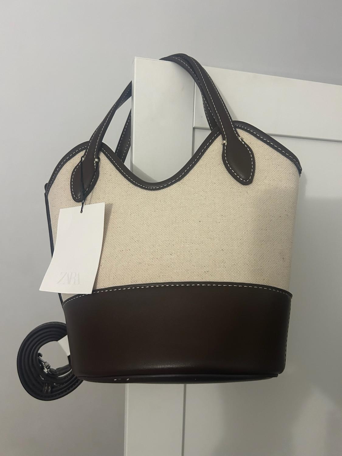 Beige fabric bag with a dark brown base and handles hanging from a white door. Label with the text ZARA.