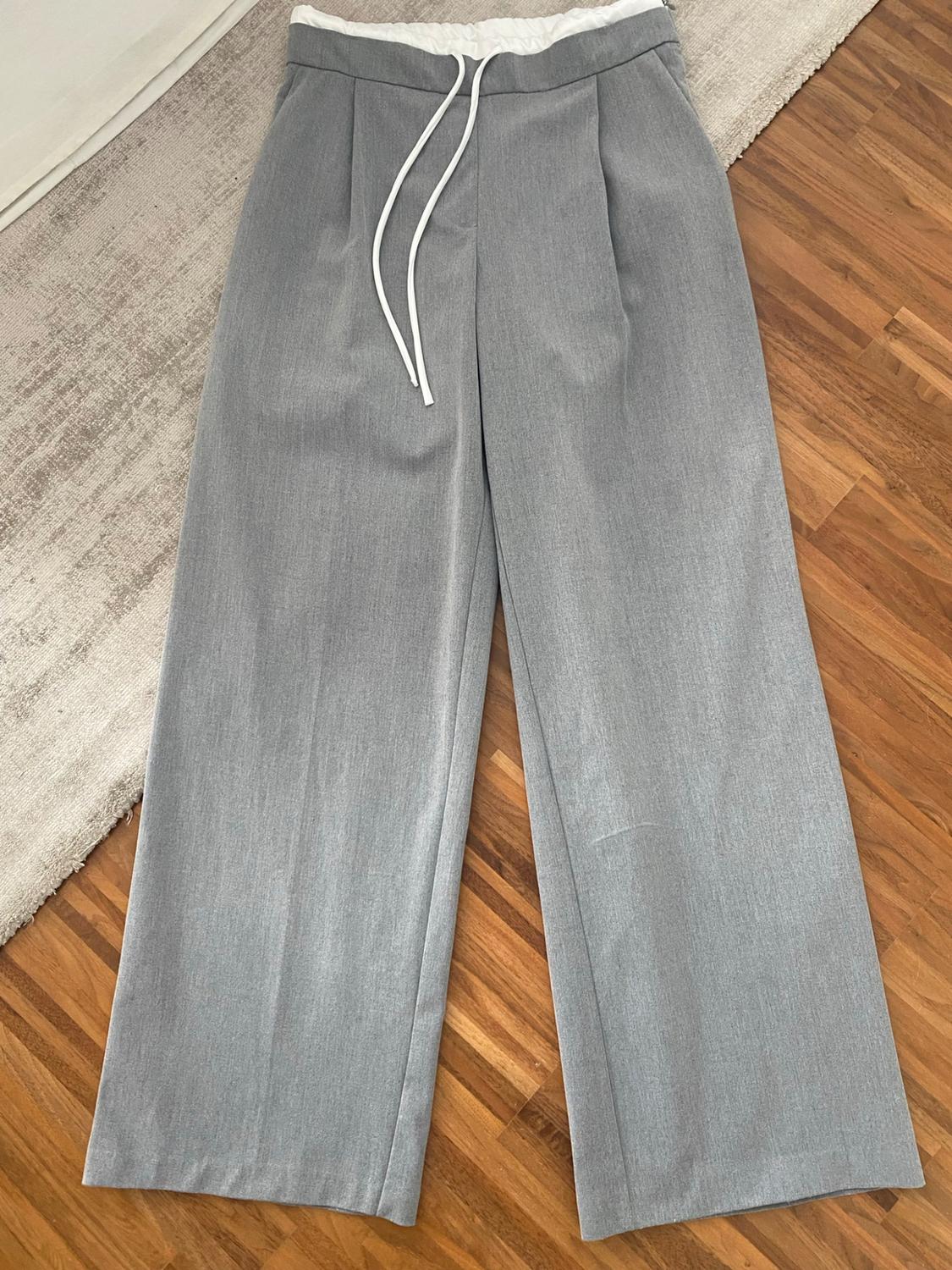 Grey wide-leg trousers with a white waistband and drawstring, laid out on a wooden floor next to a rug.