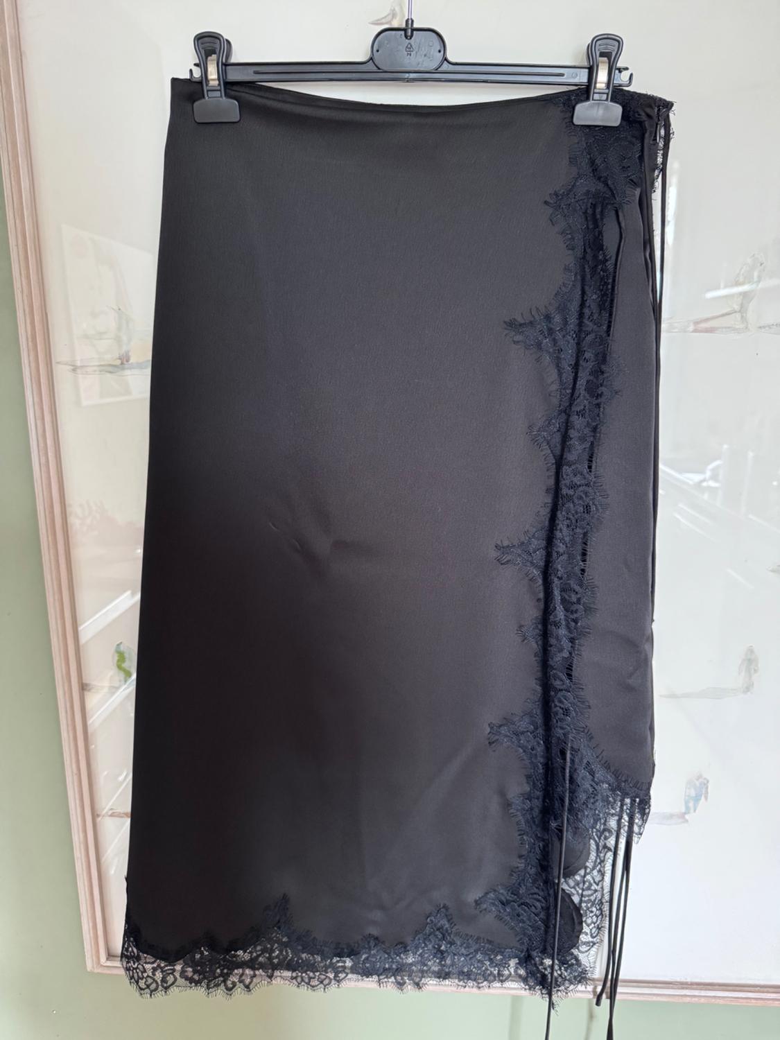Black satin skirt with lace trim, hanging on a hanger in front of a picture with sketches on a light green wall.