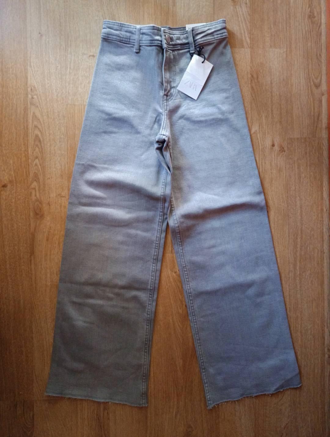 Light-grey wide-leg jeans with a frayed hem on a wooden floor. They have a label bearing the text ZARA.