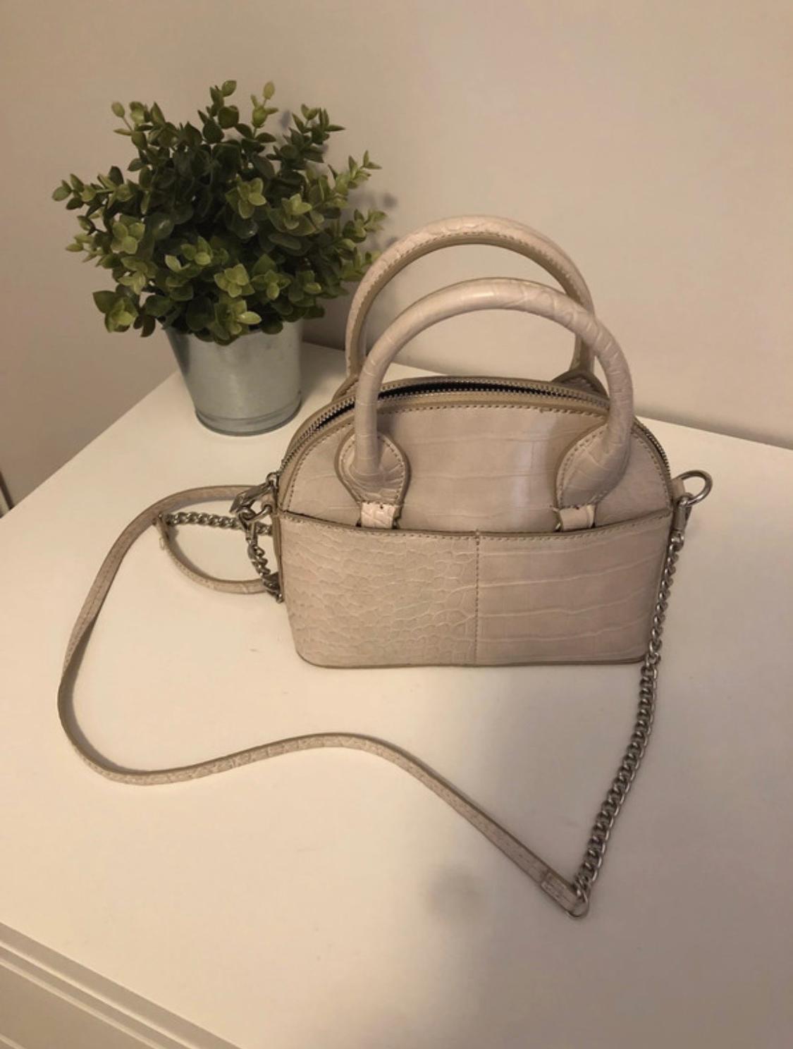 Small beige handbag with a crocodile texture, short handles and a chain strap. It rests on a white piece of furniture next to a green plant.