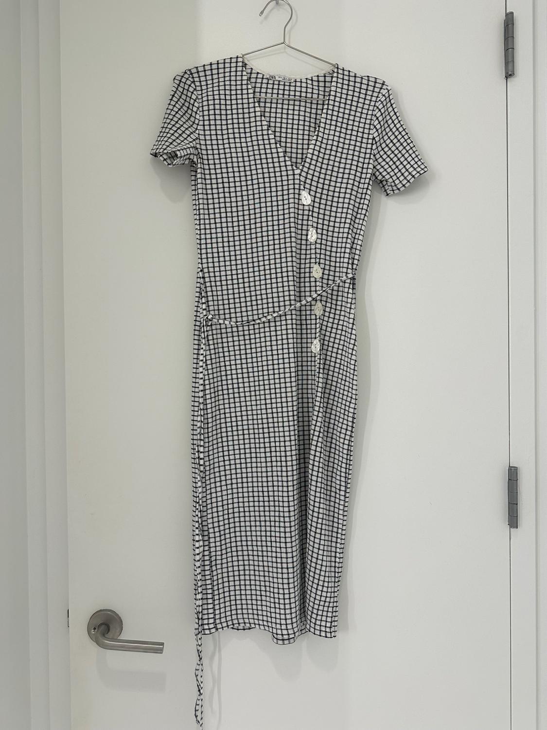 Black and white checkered midi dress with short sleeves, a V-neck, and buttons, hanging on a hanger on a white door.