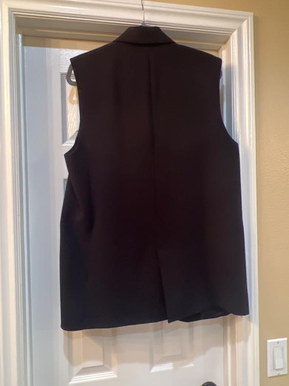 Black sleeveless vest hanging on a white door, viewed from the back. The door has a white frame and a beige wall.